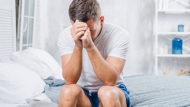 Distressed man sitting on the edge of a bed, holding his head in his hands, possibly experiencing symptoms of hormonal imbalance.